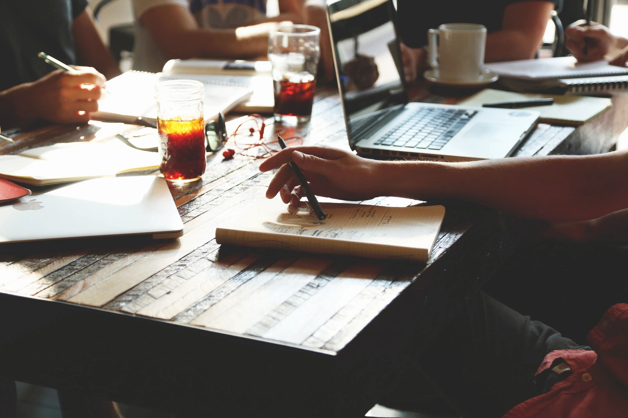 Collaborative scene of four people sitting at a table with computers, pens, notebooks, and various drinks such as coffee and soda.